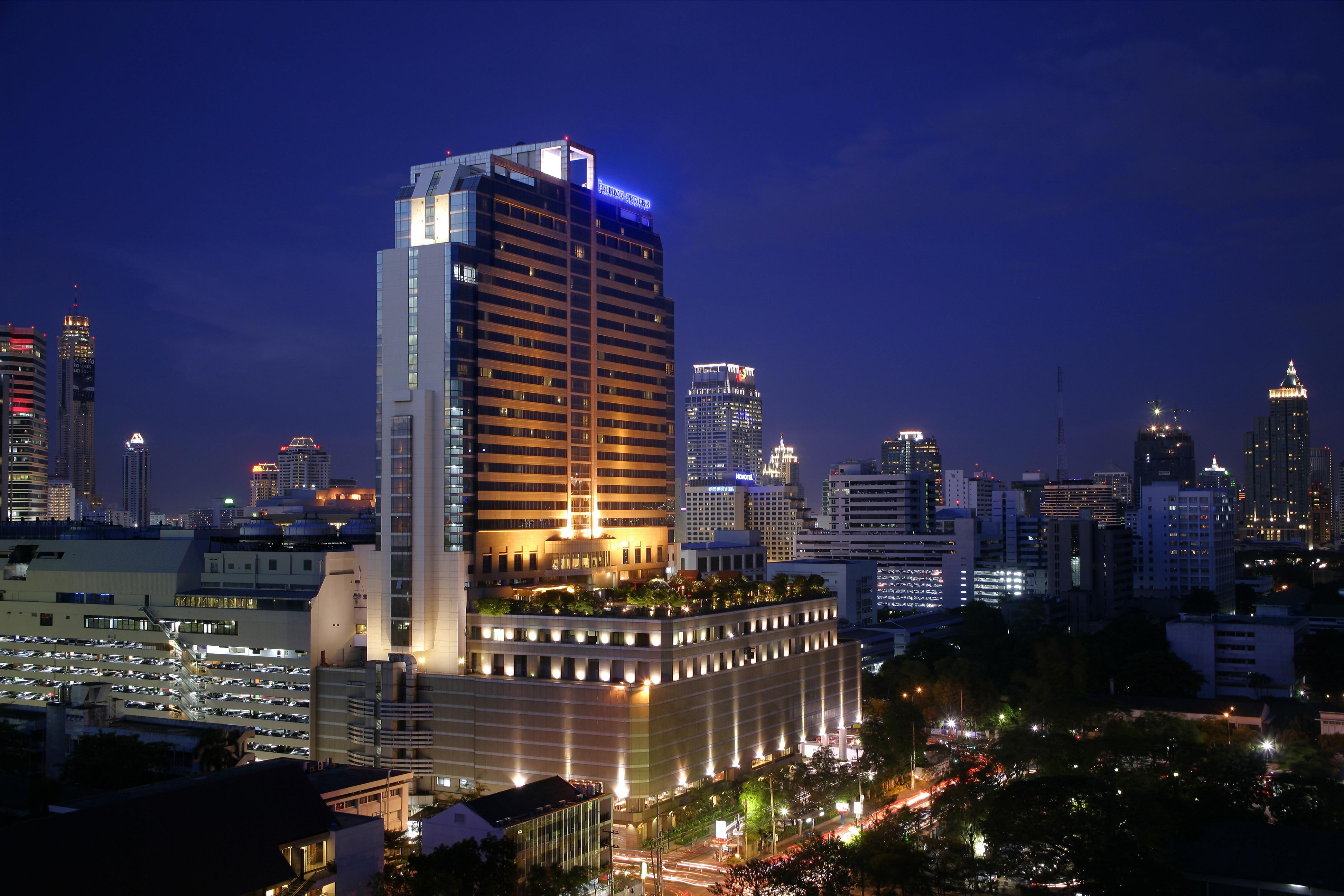 Pathumwan Princess Hotel - Sha Extra Plus Certified Bangkok Exterior photo The photo shows a cityscape at night, featuring a tall, modern hotel building illuminated with bright lights. The building has a sleek design and is surrounded by a variety of other skyscrapers and urban structures. The street below is bustling with 