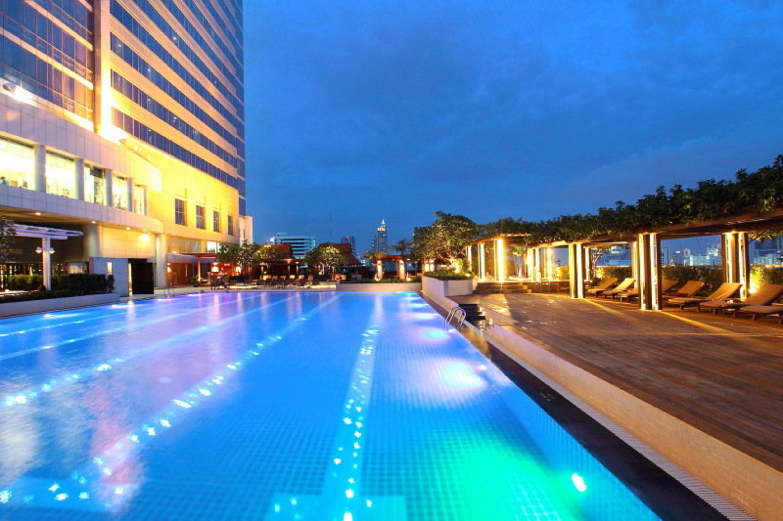 Pathumwan Princess Hotel - Sha Extra Plus Certified Bangkok Exterior photo The photo showcases a luxurious swimming pool at dusk. The pool is illuminated with colorful lights that create a vibrant atmosphere. Surrounding the pool are lounge areas with wooden decking and lounge chairs, providing a relaxing space for guests. 