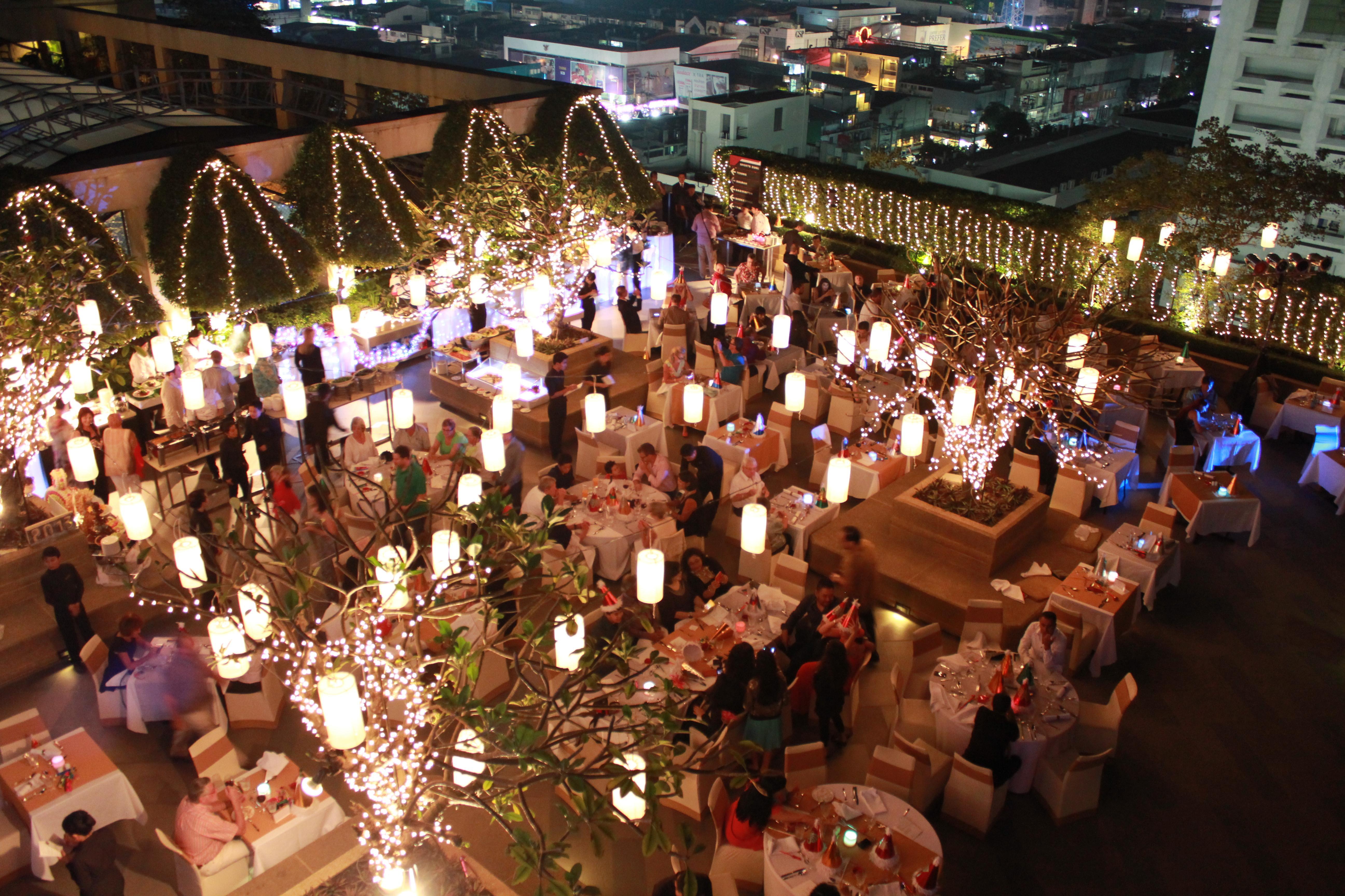 Pathumwan Princess Hotel - Sha Extra Plus Certified Bangkok Exterior photo The image depicts a spacious outdoor dining area at night, filled with tables arranged for guests. The setting is illuminated by warm, inviting lights, including hanging lanterns and fairy lights draped across trees. Many diners are visible, enjoying