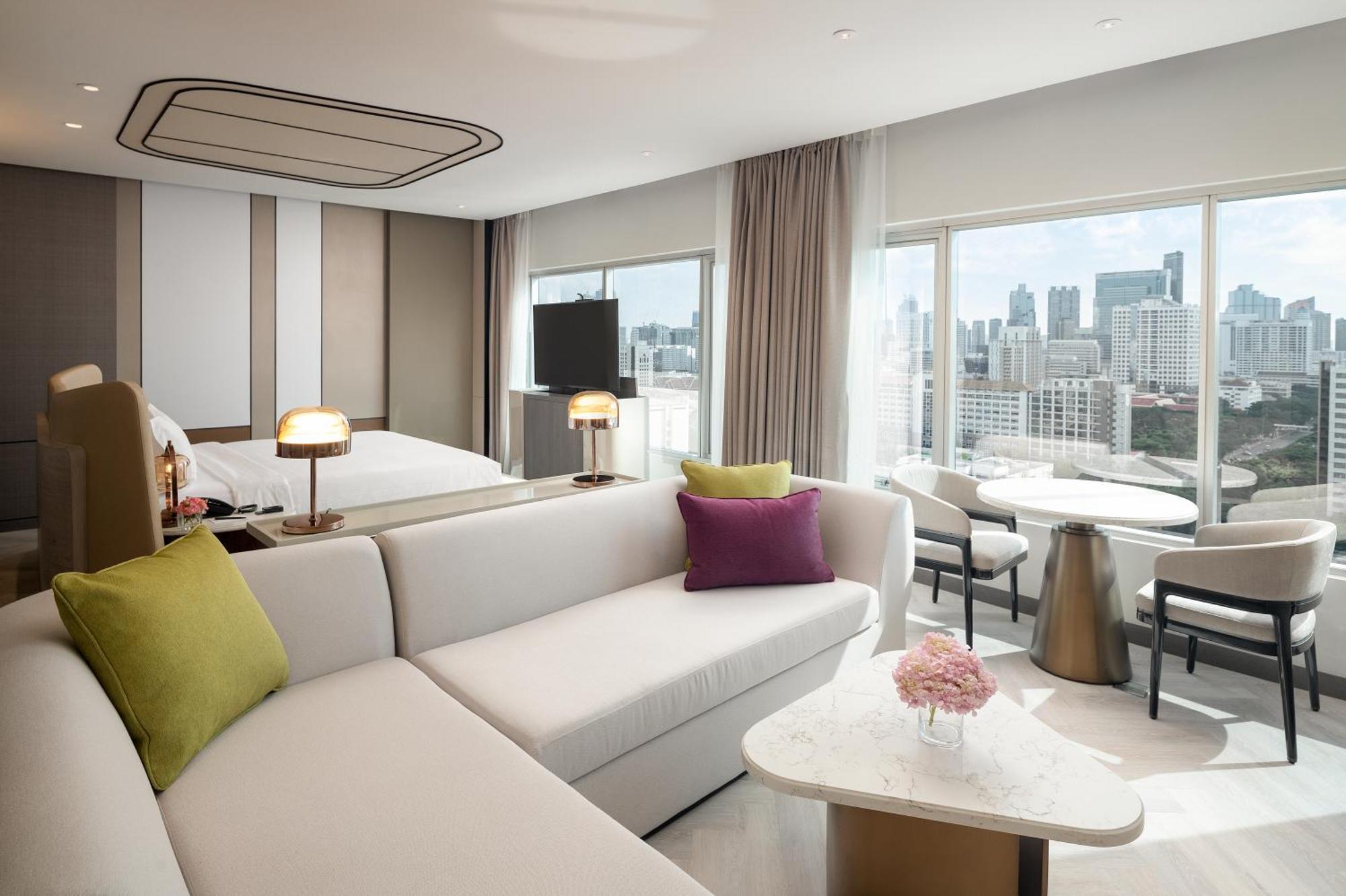 Pathumwan Princess Hotel - Sha Extra Plus Certified Bangkok Exterior photo The photo showcases a modern hotel room featuring a stylish interior design. There is a large, comfortable seating area with a white sofa adorned with colorful cushions in green and purple. Nearby, a small round table with a floral centerpiece stands