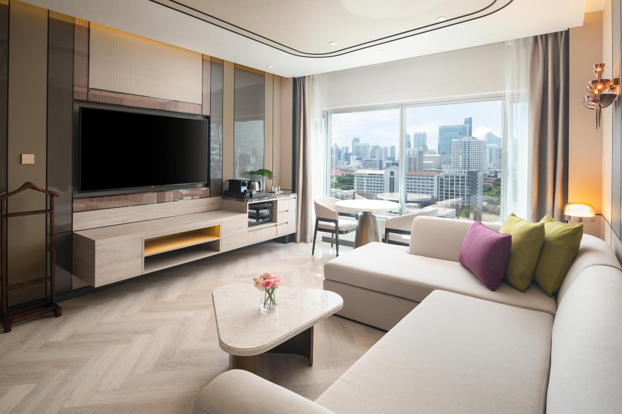 Pathumwan Princess Hotel - Sha Extra Plus Certified Bangkok Exterior photo The photo shows a modern living room with a large window offering a view of a city skyline. The room features a light-colored sofa in an L-shape, complemented by colorful cushions in shades of green and purple. A sleek coffee table sits in front of t