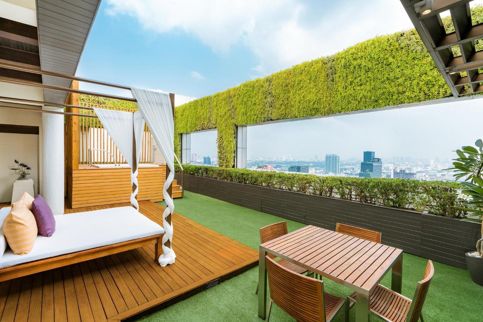 Pathumwan Princess Hotel - Sha Extra Plus Certified Bangkok Room photo The photo displays a modern rooftop terrace with a lush, green wall and panoramic views of a city skyline. The area features a wooden deck with a white, draped lounging bed, creating a relaxing space. In addition, there’s a small dining table with se