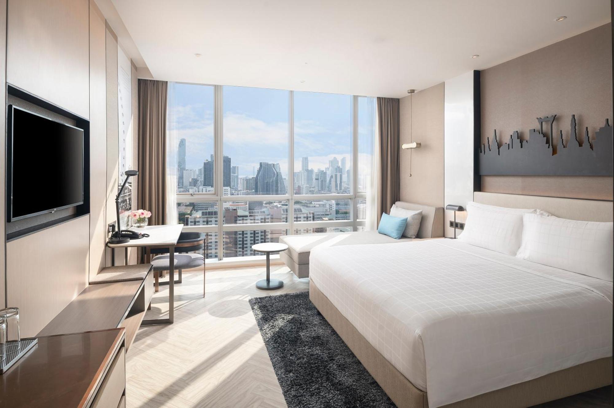 Pathumwan Princess Hotel - Sha Extra Plus Certified Bangkok Exterior photo The photo shows a modern hotel room with large windows that provide a view of the city skyline. The room features a neatly made king-sized bed, a bedside table, and a chair in the corner. There's a desk with a lamp and a flat-screen TV mounted on the